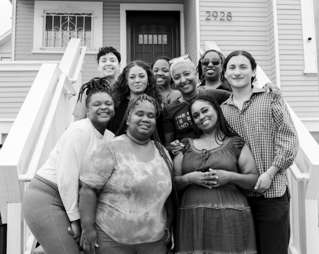 Nine people embrace each other and smile as they pose for a photo on the front porch of a house.